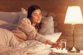 Beautiful young woman taking glass of water from bedside table in evening�