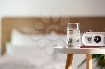 Glass of water and alarm clock on table in bedroom�