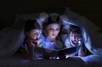 Woman and her little children reading book under blanket at night�