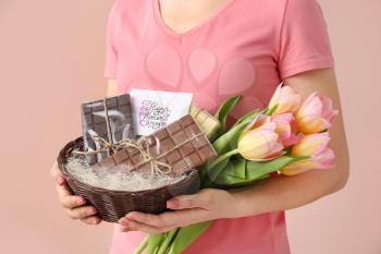 Woman holding basket with chocolate and flowers for Mother's Day on color background�