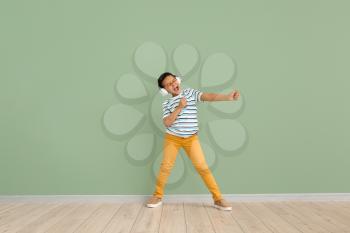 Little African-American boy listening to music and dancing against color wall�