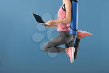 Young woman with yoga mat and laptop jumping on color background. Concept of balance between rest and work�