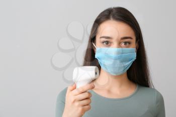 Young woman with medical mask and infrared thermometer against grey background�