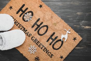 Door mat with Christmas greeting and slippers on wooden background�