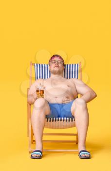 Overweight man with glass of beer sitting on beach chair against color background. Weight loss concept�