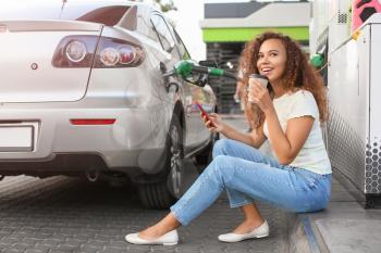 African-American woman with mobile phone drinking coffee while filling up car tank at gas station�