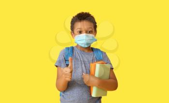Little African-American schoolboy in medical mask showing thumb-up gesture on color background. Coronavirus epidemic�
