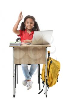 African-American schoolgirl with laptop and raised hand sitting at school desk against white background�