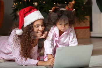 Cute African-American girl and her mother with laptop at home on Christmas eve�