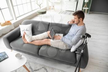 Young man with broken leg using laptop while talking by phone at home�
