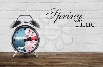 Alarm clock on table against white brick background with text SPRING TIME. Concept of time change�