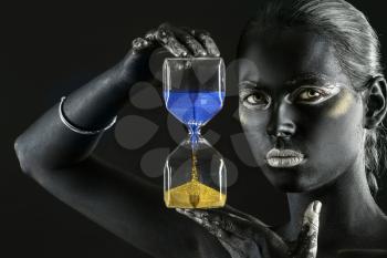 Young Ukrainian woman with black paint on her body holding hourglass against dark background�