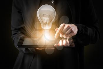 Businesswoman with tablet computer and glowing light bulb on dark background, closeup�