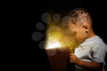 Little African-American boy reading magic book on dark background�