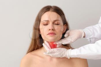Endocrinologist examining young woman with thyroid gland problem against light background 