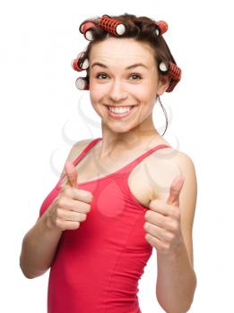 Young woman dressed in red is showing thumb up gesture using both hands while wearing hair-rollers, isolated over white