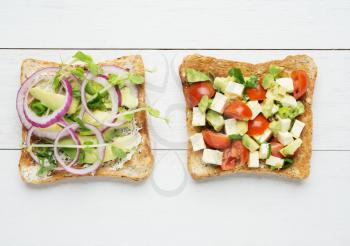 Two avocado toasts with mashed avocado and onion,sliced avocado ,tomato, snow pea sprouts and goat cheese on white wooden background
