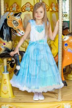 Blond little girl in blue dress standing near the carousel