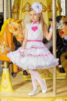 Little beautiful girl in pink and white dress standing on the carousel