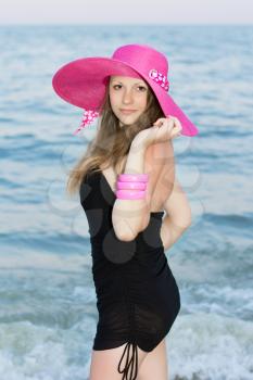 Attractive young lady wearing short black dress and pink hat posing on the beach
