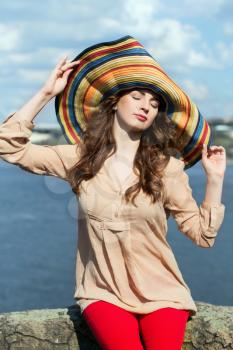 Young brunette in colorful hat sitting on a rock by the river with closed eyes