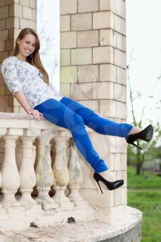 Smiling young blonde in blue pants sitting on ancient handrail