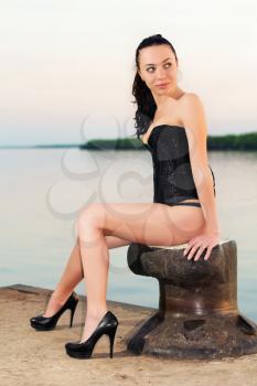 Attractive brunette in black corset and shoes sitting on the pier