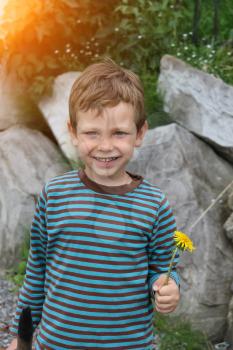 Smiling boy with dandelion in front of big rocks in sunlight