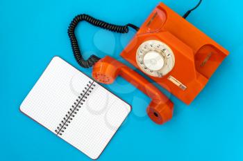  Vintage orange telephone and blank notebook on blue background, top view, pick up the phone