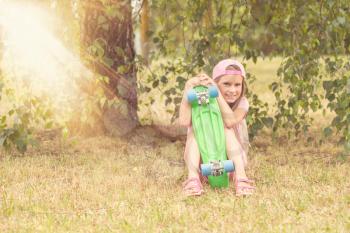 Little girl child with skate board sits under tree at summer park