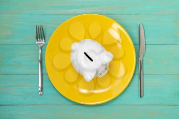 Savings consumer concept. Piggy bank on the yellow plate with fork and knife.