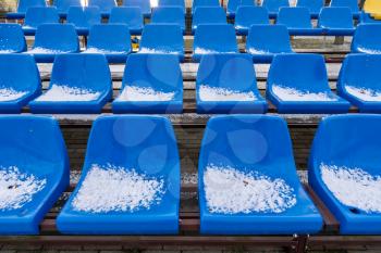 Stadium seats in the snow in winter. Snow-covered blue plastic seats at the small stadium in winter. Snowfall interferes with sports. Uncleaned stadium.