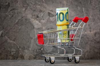 Shopping cart with rolled up Euro banknotes. Shopping costs, supermarket, finance and money concept. 