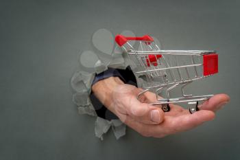 Hand with a shopping cart through a grey paper hole. Financial concept.
