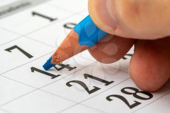 The hand of a man holding a pencil in his hand and recording his schedule on a desk calendar