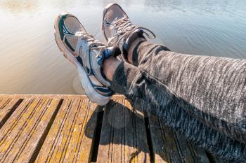 Man with crossed legs relaxing on the wooden pier. Young guy wearing sneakers sitting by the lake in summer afternoon.