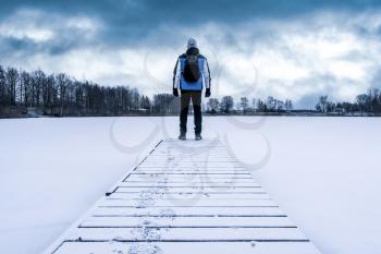 Back view of man standing on the lake wooden pier at cold winter weather