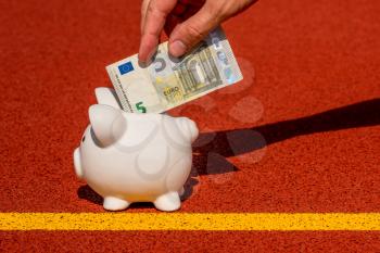 Hand putting five Euro into piggy bank. Piggy bank on the sports ground. Concept of sport fund. Money and sport.