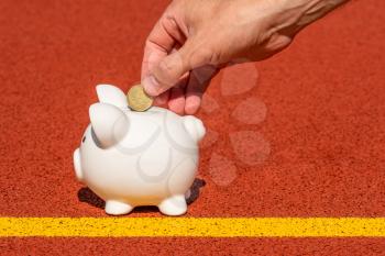 Hand putting fifty Euro cent into piggy bank. Piggy bank on the sports ground. Concept of sport fund. Money and sport.