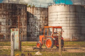 Red tractor parked near of old fuel storage tanks in an industrial area