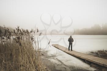 Traveller standing on a bridge in the fog. Wooden jetty on foggy day by the lakeside during winter.