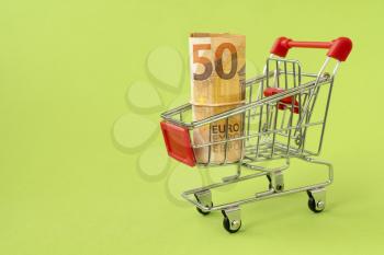 Shopping cart with roll of Euro money on green background. Copy space.