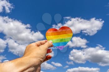 LGBT rights concept, hand holds a heart painted like a LGBT flag against sky background
