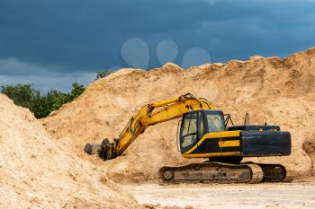 Yellow excavator during earthmoving at open pit on dark blue sky background. Construction machinery and earth-moving heavy equipment for excavation, loading.