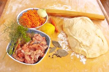 Grated carrots and a whole, onion, chunks of salmon in the pan, a lemon, three bay leaves, peppercorns, dough, rolling pin, dill and parsley on a floured board