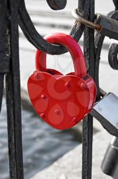 Red padlock on a heart-shaped close-up on the fence on the background of other castles, water, concrete fences