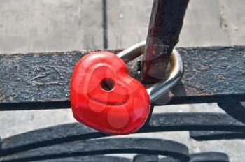 Padlock in the form of red heart on a metal fence in the background of concrete pavement