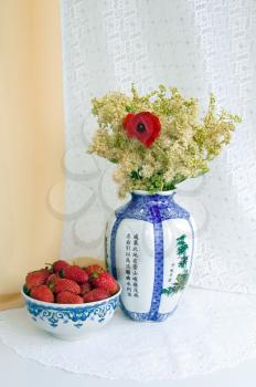 A cup of strawberries, a vase with field and garden flowers on the table with a white cloth on a background of beige and white shades