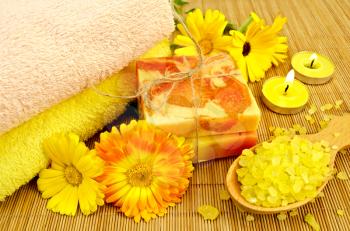 Two bars of homemade soap, towels, bath salt in wooden spoon, two candles and calendula flowers on a background of bamboo napkins