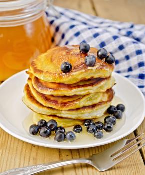 A stack of pancakes with blueberries and honey on a white plate, a jar of honey, a napkin on a background of wooden boards